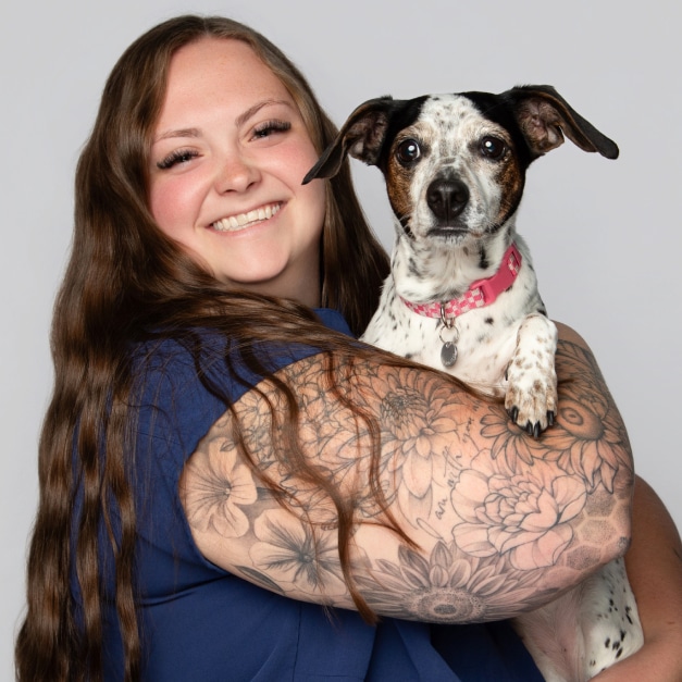 A woman with tattoos smiles while holding a small dog wearing a pink collar.
