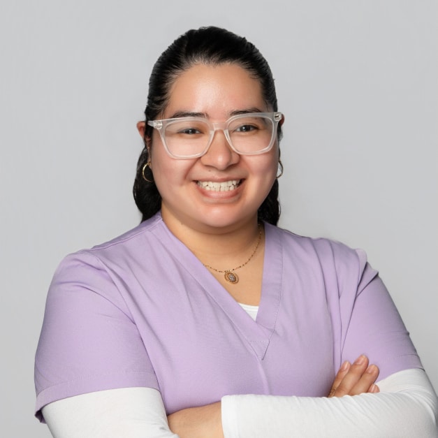 A person in a light purple scrub top smiles, wearing clear glasses and a necklace, with arms crossed.