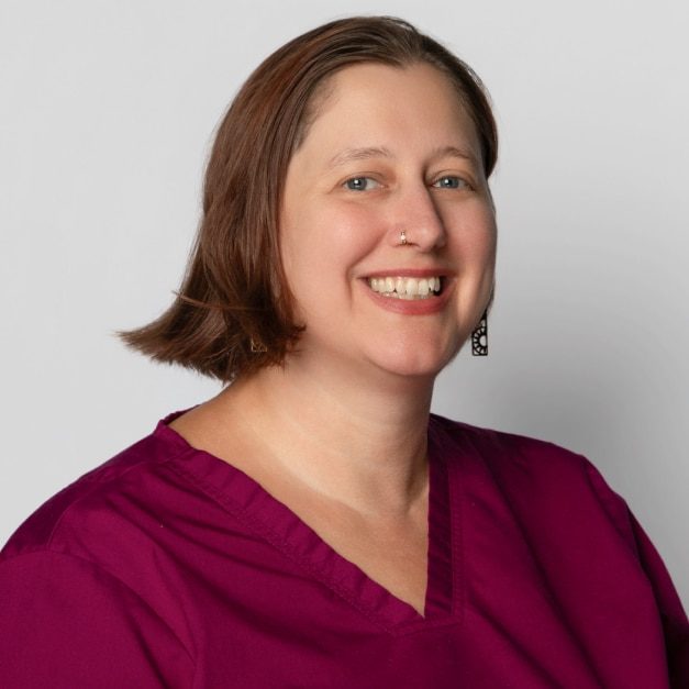 A person with short brown hair, wearing a nose ring and a magenta shirt, smiles while looking at the camera.